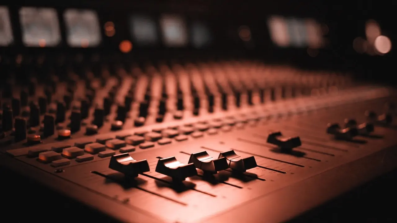 Close-up of a professional mixing console's faders washed in coral stage light, meter bridge glowing softly behind